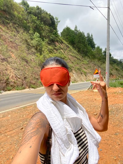 A woman with dark hair is taking a selfie while wearing an orange eye mask. She is standing outdoors on a reddish-brown dirt shoulder next to a paved road that curves to the left. The road has yellow dashed lines and a pedestrian crossing marked with yellow stripes. To the left of the road, a steep, vegetated hillside rises. To the right, a utility pole with electrical wires runs vertically. A triangular yellow traffic sign with a black symbol and the text "500M" is visible on the right side of the road. The woman is wearing a white scarf draped around her neck and shoulders and a striped top. She has tattoos on her left arm and forearm. Her right hand is raised, with two fingers extended. The sky is overcast. The environment appears to be rural, possibly in or near Phan Son, Vietnam, given the characteristic tropical vegetation and roadside infrastructure.