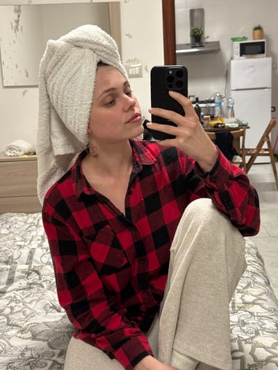The image depicts an indoor scene within the city of Ercolano, Italy. A young adult female is seated on a bed, appearing to take a selfie. Her head is wrapped in a white terry cloth towel, and she wears a red and black plaid button-up shirt over light beige knitted sweatpants. She holds a black smartphone with a multi-lens camera array in her right hand, positioned in front of her face as she looks towards the screen. Damp strands of hair are visible near her neck.

The bed she sits on is covered with a white and gray/beige patterned duvet featuring ornate designs. To the left, a light-colored wooden dresser with two visible drawers holds a neatly folded white towel and other small items. A rectangular mirror is mounted on the wall above the dresser, reflecting parts of the room and showing faint decorative patterns of leaves or flowers on its surface or the wall behind it.

The background transitions to a kitchenette area. A dark wooden structural beam is visible. A stainless steel range hood is mounted on the wall, with a small green potted plant placed on top. To its right, a white mini-refrigerator is visible, with a white microwave oven resting on top. A green bowl and another small potted plant are on the microwave. A wooden dining table cluttered with items, including water bottles and containers, is partially visible in the middle ground, with a wooden folding chair situated behind it. The walls are light-colored, and the floor appears to be tiled in a light gray or beige.