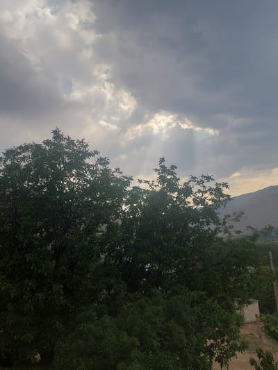 The image depicts a landscape scene with a dramatic sky. Heavy, dark grey clouds dominate the upper portion of the frame, but shafts of light break through in several areas, illuminating patches of the sky and casting rays downward. In the foreground, a large, dense tree with dark green foliage is prominently featured, its branches reaching towards the sky. Beyond the tree, a hazy mountain range is visible in the distance, with the muted tones of the sky and landscape suggesting either dawn, dusk, or overcast conditions. A small, light-colored building is partially visible on the right side of the image, nestled among greenery and a dirt path. The overall impression is one of a natural landscape under a dynamic sky, potentially indicating a change in weather. The location is identified as the city of Areni, Armenia, which is known for its mountainous terrain and vineyard regions.