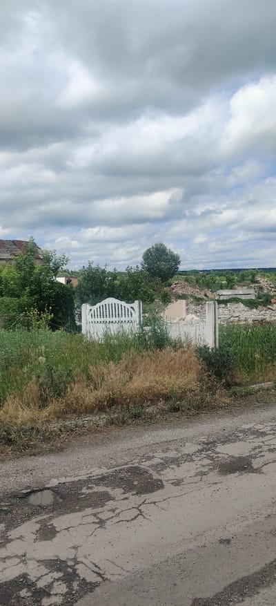In Kolomak, Ukraine, a cracked asphalt road occupies the foreground of the image. Beyond the road, a white, ornate gate stands amidst overgrown grass and weeds, with a broken wall and rubble visible behind it. In the background, there are trees, remnants of a damaged building with a dilapidated roof, and a landscape under a cloudy sky.