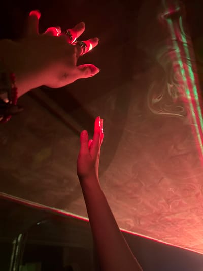 Two hands are reaching out toward a textured wall. The wall is illuminated with swirling red and white light, accented by green horizontal stripes at the top. The left hand is adorned with multiple rings and a small tattoo. A bracelet is visible at the wrist. The fingers of both hands are catching the red light, creating a glowing effect. A hint of furniture is visible in the lower right corner.