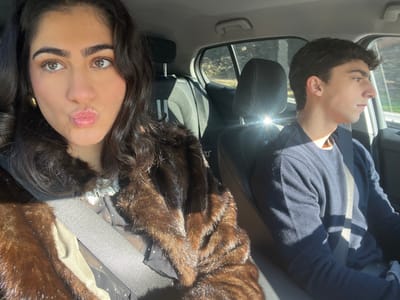 This image depicts two individuals inside a vehicle, located in Caminha, Portugal. In the foreground, a female occupies the front passenger seat, facing the camera for a selfie. She has dark, wavy hair and light brown eyes, with her lips pursed. She wears a dark brown, possibly fur, coat over a dark top with white polka dots and a lace collar. A golden hoop earring is visible on her left ear. A gray seatbelt crosses her chest. Behind her, in the rear passenger seat, a male is visible in profile, looking out the right-side window. He has short, dark, curly hair and wears a dark blue long-sleeved sweater, also secured by a gray seatbelt. Strong sunlight enters the vehicle from the right, creating prominent shadows and lens flares. The exterior background visible through the car windows includes trees and a road, indicating an outdoor, daytime setting.
