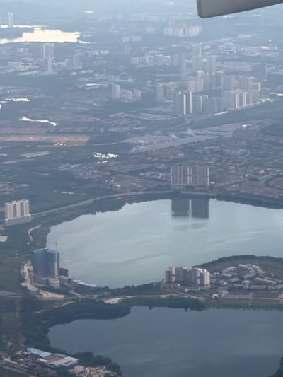 This aerial photograph captures an expansive view of a city, identifiable as Subang Jaya City Council, Malaysia. The scene is dominated by a large, calm body of water in the foreground, likely a lake or reservoir, which reflects the surrounding urban landscape. Across the water, and extending into the background, are numerous high-rise residential buildings, characterized by their uniform design and clustered arrangement. These buildings suggest a densely populated urban area.
On the western side of the lake, a construction site is visible, with a tall building under development, indicated by scaffolding and construction materials. Further to the north, on the opposite side of the larger lake, a cluster of mid-rise apartment buildings sits on a hill, overlooking the water. In the distance, more residential blocks and other urban structures are discernible, shrouded in a light haze, typical of atmospheric conditions in such environments.
The overall impression is one of a modern, rapidly developing urban center with significant residential infrastructure integrated with natural water features. The photograph appears to have been taken from a high vantage point, possibly an aircraft, given the angle and perspective. There are no visible individuals or specific activities being depicted, focusing solely on the built environment and its relationship with the landscape.