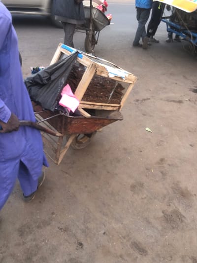 The image displays an outdoor urban scene in Tukur Tukur, Nigeria. In the foreground, a person clad in a light purple, long-sleeved garment is partially visible from the left rear, grasping the dark handle of a single-wheeled utility cart. The cart, exhibiting rust-colored metal and a wooden frame, carries a wooden display case with transparent panels. Inside the case, multiple dark, elongated items are arranged. A black plastic bag and a folded pink cloth object rest on the cart next to the display case. In the mid-ground, another individual, wearing a dark jacket and dark pants, stands near a wheelbarrow. Additional people and parts of other carts are visible in the background, along with a portion of a light-colored vehicle on the far left. The ground consists of a dusty, light grey-brown compacted surface. A single green leaf lies on the ground in the mid-right.