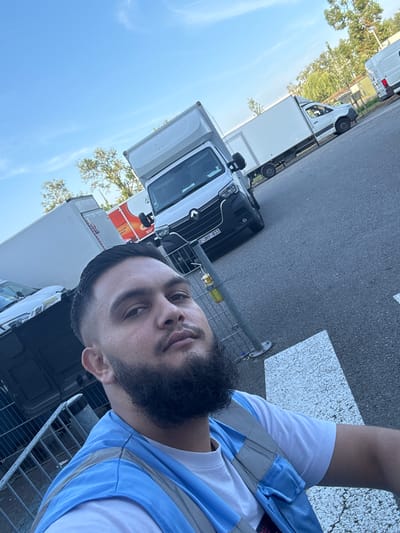 The image captures a close-up selfie of a man with dark hair and a beard, wearing a blue and gray safety vest over a white t-shirt. He is standing in an outdoor area with a paved surface, likely a parking lot or street, marked with white lines. In the background, several white trucks are visible, some with cargo boxes. One truck in the foreground has a license plate that reads "2-JHY-833". Trees and a fence are also part of the background. The sky is clear and blue with some wispy clouds. The location is identified as Boom, Belgium.