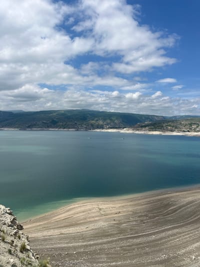The image shows a landscape with a body of water, potentially a lake or reservoir, alongside a broad, exposed shoreline that appears to be rocky and sandy. The opposite side of the water features cliffs and hills, with some development at the top of the cliffs. The sky is partially cloudy. The scene could be near Chirkey, Russia, given the likely landscape context. The image does not have people present.
