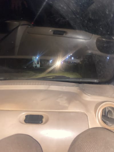 The image depicts a nighttime scene observed from the interior of a stationary vehicle in Chilаn, Uzbekistan. The foreground shows the beige dashboard of the vehicle, including a dark rectangular component embedded in its surface and the base of dark-colored windshield wipers. The top edges of light-colored fabric car seats are visible at the bottom of the frame. Through the windshield, an exterior environment at night is visible, characterized by multiple artificial light sources. A prominent, bright white light source, slightly right of center, generates significant lens flare. To its left, a vertical, illuminated structure with white and green elements is discernible, consistent with an outdoor service or vending unit. Fainter blue and white lights are scattered across the mid-ground and background, indicating a developed area. The ground outside the vehicle appears dark. The windshield exhibits reflections and potential smudges, affecting clarity.