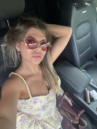 A young woman with blonde hair, wearing sunglasses and a light-colored floral dress, lies on the leather seats of a car. She is looking up at the camera with a relaxed expression. A small cross necklace is visible around her neck. The interior of the car is dark, with black leather seats. In the background, a purse and a bottle of water are visible. The scene appears to be set during the daytime, with bright lighting indicating good weather. The location is identified as Pastukhovo, Russia. There is text on a pack of cigarettes visible in the background, but it is not legible.
