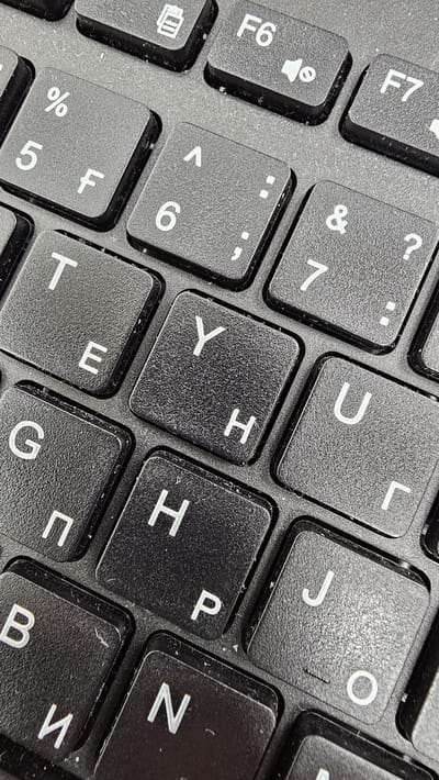 The image shows a close-up, high-angle view of a dirty, black keyboard. The keys are worn and dusty, with visible smudges and debris. Several keys are clearly legible, including F6, F7, numbers 5, 6, and 7, and letters including %, F, A, ., ;, &, ?, T, E, Y, H, U, Г, П, H, P, B, И, N, J, and O. The keys are arranged in a standard QWERTY layout, with some Cyrillic characters visible. The lighting suggests an indoor setting, possibly under artificial light. There are no people, actions, or interactions depicted. The image's context within Ақсай қалалық әкімдігі, Kazakhstan, is not visually apparent.