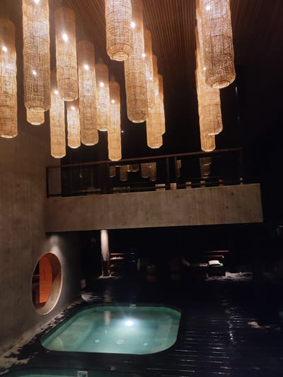 The image displays an interior space within a facility located in Mas, Indonesia, captured at night or in low light conditions. Approximately twenty cylindrical pendant light fixtures, constructed from a woven, light-colored material, hang at various heights from a dark, slatted wooden ceiling, illuminating the scene with a warm, orange light.
Below the ceiling, a concrete mezzanine or balcony, featuring a dark metal railing, extends horizontally across the middle of the frame. The area beyond this balcony is dimly lit.
On the ground level, in the foreground, an irregularly shaped, light-teal plunge pool or hot tub is visible, illuminated from within. The pool is surrounded by dark, wet wooden decking that reflects the overhead lights. A textured, light gray concrete wall forms the left boundary of the space, containing a prominent circular, wood-framed opening that leads to a dimly lit interior, possibly a sauna, with wooden paneling. In the dimly lit background of the ground level, a lounge area is discernible, including a daybed or bench with green upholstery and a white cushion. No individuals are present in the scene.