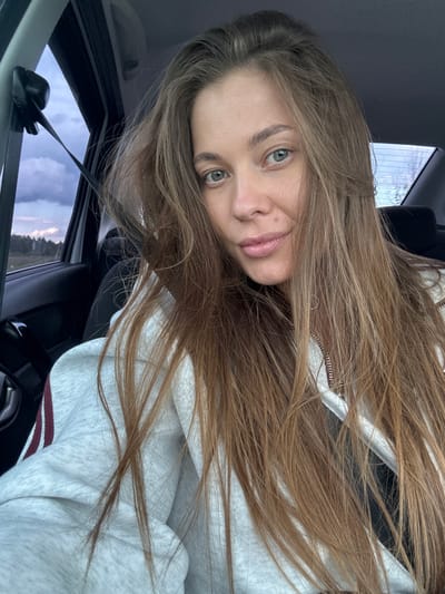 A young woman with long, light brown hair is captured in a selfie from inside a car. She is wearing a light gray hooded sweatshirt with dark red stripes on the sleeves. Her eyes are open and looking towards the camera, and her lips are slightly parted. The background shows the interior of the car, including a seatbelt, and through the window, a view of a cloudy sky over a treeline and a rural landscape. The image does not contain any elements that would suggest the location in the city of Бикбарда, Russia.