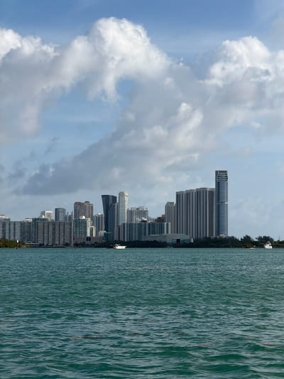 The image shows a view of Miami, Florida, United States of America. The cityscape is prominent, with numerous tall buildings and skyscrapers lining the coastline. To the left of the buildings, there are formations of white clouds in a blue sky. To the right, the scene is dominated by water with visible ripples and patterns. There is no visible indication of it being Bal Harbour Village.