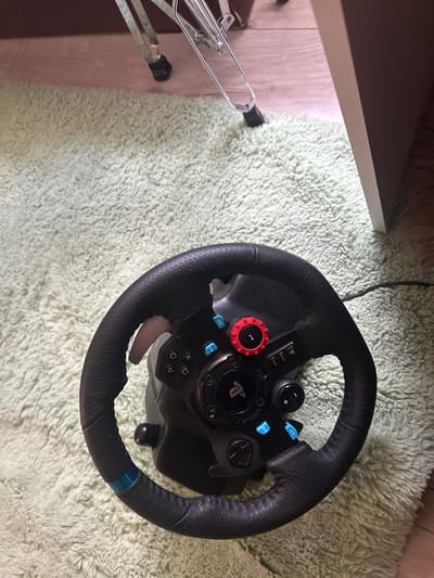 This is a top-down, close-up shot of a black gaming steering wheel resting on a light green, fluffy rug. The steering wheel has a leather-like texture and features numerous buttons and a PlayStation logo. Visible buttons include "OPTIONS," "SHARE," a red dial with an arrow, and blue-colored buttons labeled "L2" and "R2." A matte gray paddle shifter is also visible extending from behind the steering wheel. To the left of the steering wheel, a portion of a metallic object, possibly a stand or furniture leg, is visible against a light wooden floor. The background is dominated by the textured green rug, suggesting an indoor, possibly home, setting. The lighting appears to be artificial, with no distinct shadows indicating natural light sources. The image is still and focused on the steering wheel, implying it is not currently in use for gaming. The location context is Madara, Bulgaria.