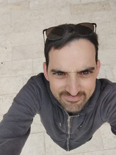 A male subject is depicted in a high-angle shot, looking upwards towards the camera, against a light-colored tiled ground in Rosh HaAyin, Israel. The individual has dark hair, a receding hairline, dark eyebrows, and green/hazel eyes. He exhibits a furrowed brow and a slight grimace or smirk, with his lips slightly parted. A short, dark beard and mustache are present. He is wearing a dark grey, zippered hoodie with the zipper partially open at the top. Black-framed sunglasses are perched on the top of his head. The background consists of light-beige, rectangular paving tiles.