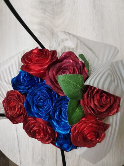 A bouquet of artificial roses, predominantly red and blue with glitter, is presented. The bouquet is partially wrapped in white paper with gray stripes, and a black stem or stand is visible. Two green artificial leaves are tucked among the roses. The background appears to be a light-colored wooden surface, possibly a table or floor. The scene is brightly lit, highlighting the sparkle of the glitter on the roses. There are no people or discernible activities taking place. The location is noted as Kuyagan, Russia.