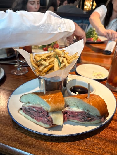 The image captures a partially eaten French dip sandwich with fries on a wooden table. The sandwich is cut in half and served with a small container of dipping sauce. Fries are served in a white paper cone that is supported by a metal holder. There are two people in the image, one is wearing a white shirt and is holding the fries, while the other is out of focus in the background. Based on visual information, the location could possibly be the "Del Frisco’s Grille" at 1221 E Southlake Blvd in Southlake, Texas.