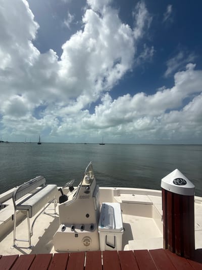The image depicts a boat docked alongside a wooden pier. The boat is predominantly white, featuring a helm with a steering wheel and a console labeled "2200". There are rod holders and a cooler mounted near the stern. The pier is constructed from red-brown wooden planks. Beyond the boat and pier, there's a view of the ocean, dotted with several sailboats in the distance. The sky is filled with numerous white clouds against a blue background. The scene likely is in Key Largo, Florida, suggested by the coastal environment.