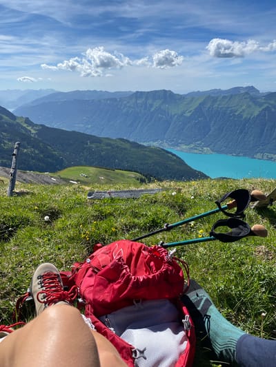 The image shows a pair of legs and a red backpack resting on a grassy slope overlooking a stunning mountain and lake landscape. The person is wearing hiking boots with red laces and green patterned socks. Trekking poles are placed beside the backpack. In the background, a vast expanse of mountains stretches towards the horizon, with a bright turquoise lake visible in the mid-distance. The sky is blue with scattered white clouds. The location is identified as Axalp, Switzerland.