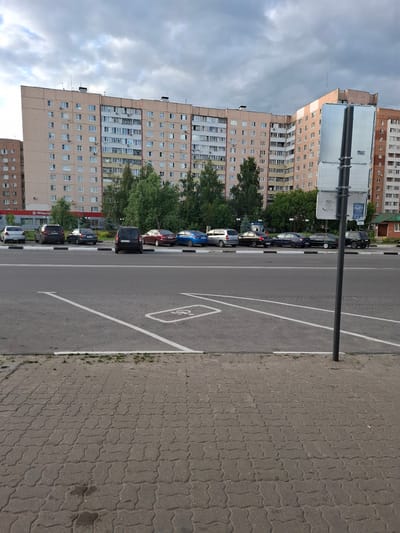 The image depicts a street scene in Меткомелино, Russia, on a cloudy day, likely in the late afternoon. The foreground features a paved walkway with interlocking bricks and a portion of an asphalt road marked with white parking lines, one of which indicates a disabled parking space. A black metal lamppost stands prominently on the walkway.
The mid-ground of the image showcases a row of parked cars along the street, including a dark SUV, several sedans, and a blue car. Beyond the cars, a line of lush green trees partially obscures a tall, multi-story residential building. This building, constructed with a mix of beige and lighter-colored facades, features numerous windows and balconies. Further in the background, another, shorter apartment building is visible.
The sky is overcast with dense, grey clouds, suggesting the possibility of rain. No people are visible in the frame, and the general atmosphere is one of an ordinary urban environment. A small blue sign with white text is partially visible on the lamppost, but the text is illegible. Another sign, partially visible on the side of a building in the background, appears to be a red circle with white text, possibly indicating a local business or service, and the word "Пятёрочка" (Pyaterochka, a Russian supermarket chain) is visible on a red sign under the building.