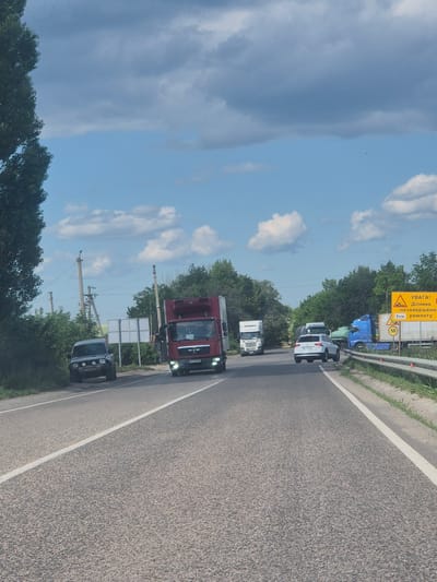 This media file depicts a traffic scene on a road in Pidhaitsi, Ukraine. The image is oriented vertically, with the sky occupying the upper left portion of the frame and the road surface filling the right side. Several vehicles are visible, predominantly trucks, lined up on the road. In the foreground, a red truck with "MAN" branding on its grille is prominent. Behind it, a white truck and a blue truck are also visible, suggesting a traffic jam or a queue. Further down the road, a white car is parked or stopped.
The setting appears to be a rural or semi-rural area, with lush green trees lining the sides of the road. The sky is partly cloudy with some areas of blue sky and patches of white clouds, indicating fair weather, possibly late morning or early afternoon given the lighting. There are some road signs visible. One prominent yellow sign has Ukrainian text and a warning symbol, likely indicating road work or a hazard. Another smaller sign shows a circular speed limit of "50". Electrical poles with wires are also present along the roadside. The overall impression is of a typical day on a highway with some traffic congestion.