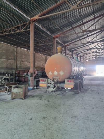 The image depicts a tanker truck parked inside a large, possibly industrial building in Ovcharovo, Bulgaria. The tanker is orange and cylindrical, resting on a truck chassis. The building has a high, corrugated metal roof supported by a metal framework. The floor is concrete. The truck appears to be stationary. The environment suggests an enclosed storage or maintenance facility.