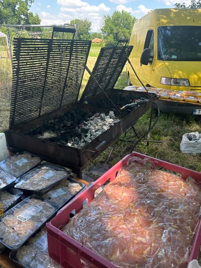 This media file depicts an outdoor barbecue scene, likely in Lovets, Bulgaria. The primary subjects are the preparations for a cookout, with a large charcoal grill taking center stage. The grill is filled with lit coals, and several metal grates are positioned above it, ready for use.

Adjacent to the grill, there are several containers filled with raw meat. One large red plastic bin is packed with thinly sliced meat, possibly chicken or pork, covered in plastic wrap. Below this, multiple pre-packaged trays of what appear to be meatballs or patties, also wrapped in plastic, are stacked neatly. The packaging shows labels with Cyrillic text, consistent with the Bulgarian location. A yellow van is visible in the background, suggesting a catering event or a large gathering.

The scene is set in a grassy area with trees and a blue sky with scattered clouds, indicating fair weather, likely during daytime. The overall atmosphere suggests anticipation and preparation for a meal. There's no visible text in English, but the product labels on the meat trays and the license plate on the van contain Bulgarian characters.