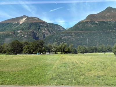 This image depicts a pastoral scene with a vast green meadow in the foreground. In the midground, a line of trees borders the meadow, and behind the trees, there are a few structures including what appear to be small buildings or sheds, white fencing, and a turquoise vehicle. A utility pole is also visible in the field. The background is dominated by a majestic mountain range, covered in dense green foliage, with rocky, exposed sections on the upper slopes and summit. The sky above is a clear blue with thin, wispy clouds. The overall impression is one of a tranquil rural landscape, likely in a mountainous region. The location is identified as Erstfeld, Switzerland, which is known for its alpine surroundings. There are no people visible in the image.
