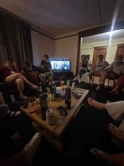 A group of eight individuals are gathered in a dimly lit living room in Harbor Springs, United States of America, with a television on in the background displaying a muted program. The room appears to be a residential space, possibly a shared apartment or house, with furniture including couches, chairs, and a large wooden coffee table.
On the coffee table, various objects are present, suggesting a casual social gathering. These include several cans of beer, a bottle of wine, a water bottle, glasses with drinks, and a bag of chips. Remotes for the television are also visible on the table. The floor is carpeted, and curtains hang on the wall to the left.
The individuals are in various states of relaxation and engagement. Some are seated, while others appear to be standing or leaning. Their attire is casual, with t-shirts, shorts, and casual dresses being common. The atmosphere seems relaxed and informal. The lighting is predominantly from the television screen and a bright light source at the top right of the image, creating some glare. The overall scene depicts a typical social gathering of friends in a comfortable, indoor setting.