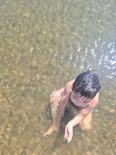 A young boy with dark hair is standing in shallow, clear water with a rocky bottom. The sun is shining brightly, creating reflections on the water's surface. The boy is looking down at his hand, which is holding a small, light-colored stone. He is wearing swim trunks, and only his upper body and arms are visible. The scene appears to be in a natural body of water, possibly a river or stream, in the city of Novoishimsky rural district, Kazakhstan.