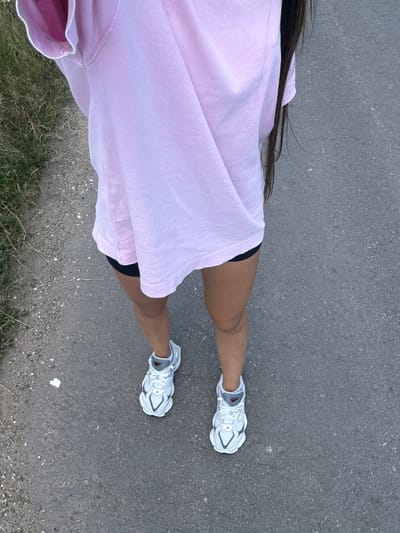 A person wearing a pink oversized t-shirt and black shorts stands on a paved surface. The person is wearing white and grey sneakers with white laces. The background shows dry grass and a grey, asphalt-like ground. The image appears to have been taken outdoors in a rural or suburban setting, possibly in Serpneve, Ukraine, given the context. The lighting suggests it is daytime, with no specific weather conditions apparent.