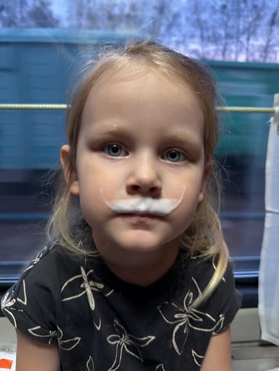 A close-up, candid portrait features a young child with fair skin, light blonde hair, and prominent blue eyes, looking directly at the camera. A distinct "milk mustache" or white foam is visible above the child's upper lip, indicating recent consumption of a beverage. The child wears a dark-colored t-shirt adorned with a repeating pattern of white floral designs. Their head is slightly tilted, and their expression appears calm and engaging.
The background, viewed through a window, is softly blurred, creating a shallow depth of field. This exterior scene reveals what appears to be a wintry or early spring landscape with light-colored, bare trees or bushes against a bright, possibly overcast sky, suggesting it is daytime. Interior elements like a window frame, a white curtain along the top edge, and a vertical light-colored support bar are also discernible, suggesting the child is indoors, potentially within a vehicle such as a train or bus. The overall mood is spontaneous and playful. No visible text is present in the image. The location context for this scene is Michurinsk, Russia.