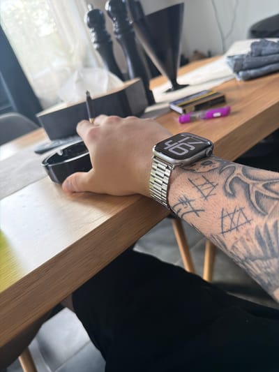 The image depicts a close-up view of a person's arm and hand resting on a wooden table. The person is wearing a smartwatch with a metallic band, and the screen displays the time "46". The arm has tattoos with geometric designs. In the background, there is a tissue box, an ashtray with a cigarette in it, and some stacked fabrics. The scene appears to be indoors, possibly in a home or office setting, with natural light filtering in from a window. The provided location information "Lozno, Bulgaria" is not visually supported by any elements within the image itself.
