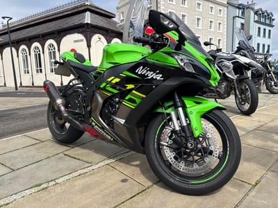 A bright green and black Kawasaki Ninja motorcycle, model 1000, is prominently displayed in the foreground, parked on light grey paving slabs. The motorcycle features "Ninja" branding in white script on its fairing, "Kawasaki Racing Team" on the lower fairing, and an Akrapovic exhaust system. Its front wheel is equipped with Brembo brakes and Bridgestone tires with green rim accents. Behind the Ninja, to the right, a white and silver BMW GS adventure motorcycle is visible, with a third, darker motorcycle parked further back.

The scene is set in Douglas, Isle of Man. To the left and behind the Kawasaki, a historic building with light-colored walls, dark ornate trim, and multiple arched windows is visible, featuring a dark roof with an intricate decorative railing. A street sign "MARKET HILL" is affixed to this building, identifying it as the Market Hall area. In the background, to the right of the motorcycles, are multi-story buildings: one is a light grey structure with numerous windows (partially obscured, but suggesting "S HOTEL"), and another further right is a light blue-grey building with multiple windows. The ground consists of concrete paving slabs interspersed with white gravel borders and small patches of grass. The lighting suggests a bright, clear day, casting subtle shadows. No people are visible in the scene.