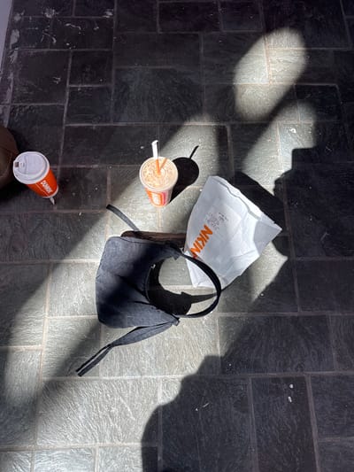 The image presents an overhead view of several items placed on a dark, textured tiled floor, strongly illuminated by sunlight. The floor consists of rectangular, dark grey tiles with a slightly uneven, possibly slate-like surface, and shows some light-colored scuff marks or debris in various areas.
The main subjects include a white paper bag, a black handbag, an iced coffee cup, and a hot coffee cup. The white paper bag is situated near the center of the frame, displaying part of the orange and white "Dunkin'" logo, specifically the letters "NKIN," and a label that clearly states "Drive thru" followed by the number "140224" and other small, illegible text. Adjacent to the bag is a black fabric handbag with a soft, slightly gathered texture and a short strap.
To the left of the paper bag, an iced coffee cup with a straw is visible, filled with a light brown beverage and featuring the "Dunkin'" logo. Towards the bottom right, an orange hot coffee cup with a white lid, also branded with "Dunkin'," is placed on the floor.
Strong, angular shadows are cast across the floor by unseen structures or objects, indicating bright, direct sunlight, suggesting a sunny day, likely mid-morning or afternoon. The arrangement of the items implies a temporary stop or a moment of rest, possibly after acquiring food and drinks from Dunkin'. The scene is likely set in an indoor or covered outdoor area with a tiled floor, located in the Town of Red Hook, United States of America. No individuals are present in the image.