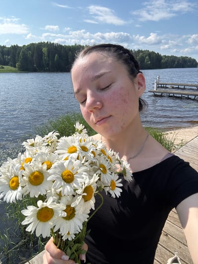 This is a vertical, close-up, outdoor, selfie-style shot of a young person holding a bouquet of daisies. The subject is positioned on the right side of the frame, their face partially visible with eyes closed, head tilted back slightly. Their skin shows some redness, particularly on the cheeks and forehead. They are wearing a black t-shirt and a silver chain necklace. The bouquet of daisies is held in the lower right corner of the frame, obscuring part of the subject's chest and arm.
The background features a body of water, likely a lake, with a treeline on the far side. A wooden dock or pier is visible in the upper right portion of the image, with some wooden structures extending into the water. The sky above is blue with scattered white clouds. The lighting suggests it is daytime, likely late afternoon or early evening, given the warm glow and shadows. The overall mood appears peaceful and serene, perhaps evoking a sense of enjoying nature. The location is identified as Pozdyshki, Russia, which aligns with the naturalistic scenery. There is no visible text in the image.