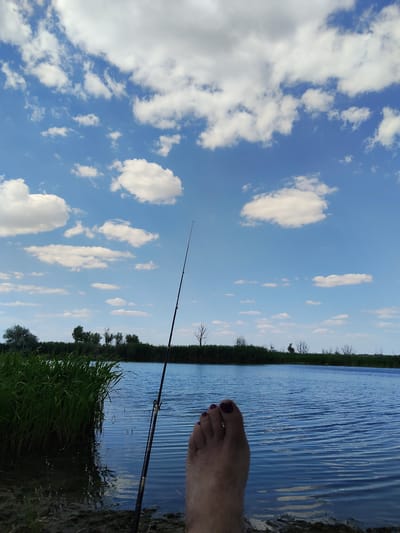 The image depicts a serene outdoor scene by a body of water under a bright blue sky dotted with fluffy white clouds. A fishing rod stands upright in the foreground, positioned to cast into the water. In the lower right corner of the frame, a person's bare foot is visible, with toenails painted a deep red. The foot rests on the muddy bank of the water. The water itself is a deep blue, with gentle ripples suggesting a light breeze. Along the far bank, a dense line of tall green reeds and various trees creates a natural horizon. The overall impression is one of peaceful relaxation and connection with nature. The scene appears to be set during the daytime, with clear skies and pleasant weather conditions, ideal for outdoor activities like fishing. There is no visible text in the image.