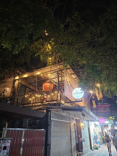 The image captures a vibrant street scene at night in Ban Klong Haeng, Thailand. The main subject is a multi-story establishment with an open-air, industrial-chic design, illuminated by strings of warm, decorative lights. This establishment appears to be a bar and restaurant, as indicated by prominent signage including "THE BLUE MARE Restaurant&Bar," "Casablanca BAR & RESTAURANT," and "COFFEE." Other visible text includes "Bar Dining," "ROOF TOP," and various food and drink offerings in Thai and English, such as "Thai Food" and "European food."

The building itself is constructed with a metal frame and corrugated metal siding, with translucent fabric awnings creating covered areas. Ornate hanging lamps and a large, circular woven light fixture add to the ambiance. On the right side of the image, a closed shuttered storefront suggests that some businesses on the street may have closed for the night, while others, like the brightly lit establishment, are still open and inviting.

In the background, a bustling street can be seen with more illuminated signs and the silhouettes of people walking by, hinting at a lively nightlife atmosphere. The dense foliage of trees frames the upper left portion of the image, their leaves illuminated by the ambient light, suggesting the presence of lush greenery in the area. The time of day is clearly night, and the overall mood is energetic and inviting, characteristic of a popular tourist or entertainment district.