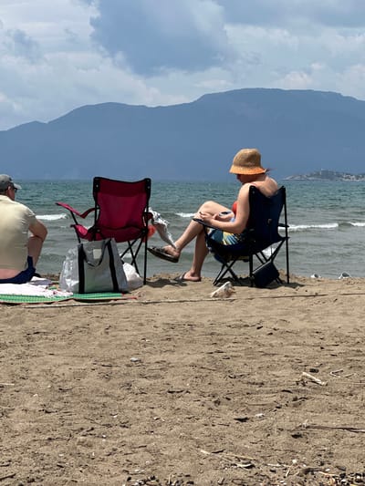 The scene captures a beach setting near Ortaca, Turkey. A woman wearing a straw hat and swimsuit sits on a blue folding chair near the water's edge. A red folding chair and a silver tote bag are nearby. The shoreline consists of sand, and the sea stretches out towards a distant mountain range under a cloudy sky. Another person is partially visible, lying on a towel.