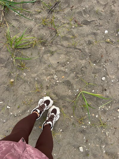 The image shows a top-down view of a person's legs and feet standing on a sandy surface covered with sparse vegetation and small pebbles. The person is wearing brown leggings and white and brown athletic shoes. The sandy ground shows some subtle footprints. The location is described as a beach in the city of Aniva, Russia. The overall environment appears to be natural and undeveloped.