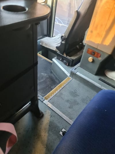 The interior of a bus is shown from a low angle. A dark seat with a pink strap is in the foreground. Beyond the seat is the floor, with a yellow and grey safety strip. A blue seat is in the upper right corner. To the left, the driver's area is visible, featuring a cracked windshield, a white cloth, and various controls. The light source appears to be from the top left of the frame.