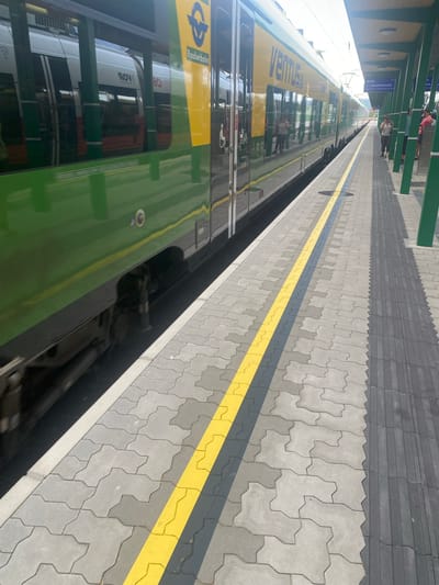This image captures a scene at the train station in Wulkaprodersdorf/Vulkaprodrštof, Austria. A green and yellow train, identified as "VENTUS", is stopped at the platform. The train's exterior is reflective, showing glimpses of the interior and people. The platform itself is paved with interlocking grey concrete blocks, with a distinct yellow and grey safety line marking the edge. A textured grey tactile paving strip runs alongside the safety line, indicating the platform's edge for visually impaired individuals. In the background, the station's covered waiting area is visible, supported by green pillars. A sign indicates a train departure for "REX Deutschkreuz". Several people are standing on the platform, waiting for or disembarking from the train. The overall impression is one of a functional and active train station environment on a bright, possibly sunny day, given the strong light and shadows.