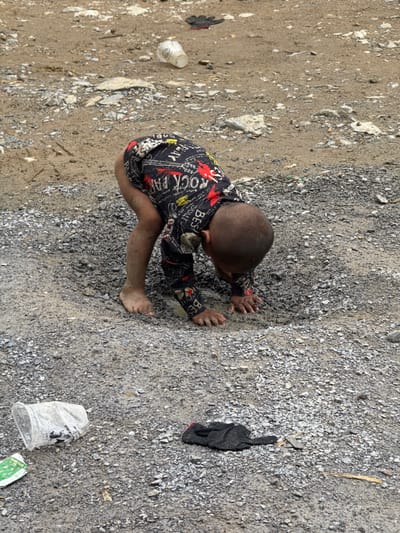 A young, bare-footed child in a black shirt with red and white printed designs is on the ground, hands outstretched, in Phố Bảng Commune, Vietnam. The ground is a mix of dirt, gravel, and various debris. A black object (possibly a glove) and a white plastic cup are on the ground near the child.