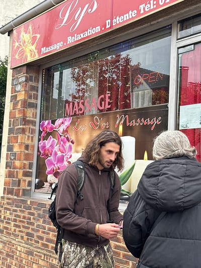 The image depicts an outdoor scene in Saint-Gratien, France, in front of a commercial establishment named "Lys." The building has a brick facade on the lower section, transitioning to an upper section with a red sign displaying "Lys" in white script, along with "Massage . Relaxation . Détente" and a telephone number. A yellow, stylized floral emblem is present on the left side of the red sign.

Two individuals are present on a sidewalk directly in front of the building's large display window.
1.  A male subject with long, dark, curly hair and a beard stands on the left. He is wearing a dark brown zippered hoodie, camouflage-patterned pants, and a black backpack. A light-colored band is visible on his right wrist. He is holding a small, light-colored object, possibly a card or paper, in his right hand and is oriented towards the second individual, appearing to engage in an interaction. His gaze is directed downwards and to the right.
2.  An older female subject is positioned with her back mostly to the camera, partially obscuring the male's right side. She has gray hair pulled back and is wearing a dark, possibly black, padded jacket. Her posture suggests she is facing the male subject.

The display window behind the individuals features large white text spelling "MASSAGE" and "Lys d'or Massage." Below this, a large image background includes vibrant pink orchids and large white candles with illuminated wicks. Reflections in the window show overhead structures and autumnal-colored trees. An illuminated "OPEN" sign is visible as a reflection in the upper right portion of the window. The overall scene suggests a casual street encounter in front of a massage and relaxation parlor.