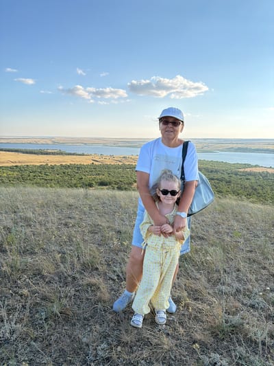 A woman and a young girl stand on a grassy hillside overlooking a body of water and a wooded area. The woman, wearing a white t-shirt, denim shorts, a white baseball cap, and sunglasses, has her arm around the girl. The girl, dressed in a yellow checkered outfit and sunglasses, is holding the woman's hands. A light blue bag is slung over the woman's shoulder. The sky above is blue with scattered white clouds. The landscape suggests a rural or natural setting, possibly near Gornyi Balyklei, Russia, given the context.