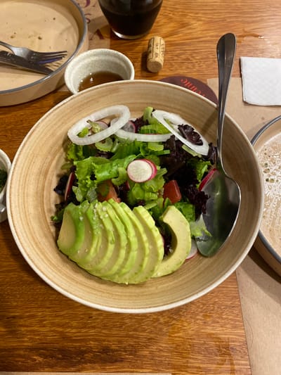 A bowl of salad with avocado slices, onion rings, radish, lettuce, and other greens is on a wooden table in Punta Hermosa, Peru, at the coordinates -12.333, -76.826. There is a silver spoon in the bowl. Next to the bowl is a partially filled glass of dark liquid, a small white plate with a fork, a small bowl with a clear liquid and a brown cork. There is also a napkin and a white plate partially filled with a clear liquid in the background. The table is brown and wooden. The bowl is beige and slightly textured. The table is also covered in a beige napkin.