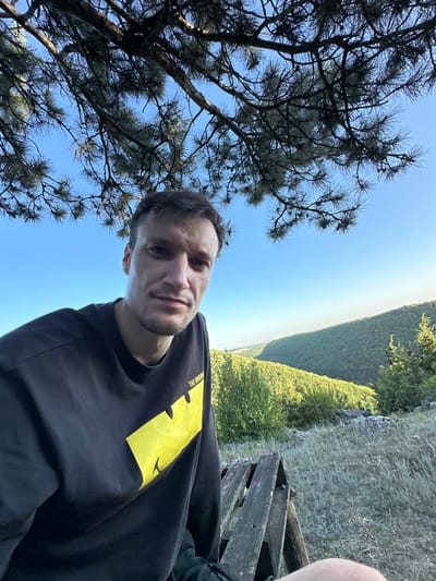 A man with short brown hair and stubble is taking a selfie outdoors, seated on a wooden structure. He is wearing a black long-sleeved shirt with a yellow graphic. The background shows a lush, green valley under a clear blue sky. The scene is illuminated by natural sunlight. The location is in the city of Pryvorottia Druhe, Ukraine.