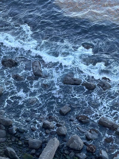 This aerial shot depicts the coastline near Staffin, United Kingdom. Rocks of varying sizes occupy the right side of the frame, bordering a turbulent ocean. White foam traces the patterns of the wave action against the dark blue water. The left side shows calmer water, transitioning from a light, sandy color to the deeper blue of the open sea.