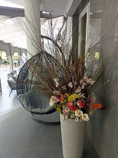 A high-angle, eye-level medium shot shows a hanging rattan chair with a gray cushion, positioned on a gray tiled patio next to a large, decorative vase filled with a floral arrangement. The vase is cylindrical with a textured, wave-like pattern and is placed in front of a dark gray stone wall. The floral arrangement features a mix of artificial flowers in various colors including pink, yellow, red, and orange, interspersed with dried branches. In the background, a white canopy is visible, supported by columns, and further beyond, a covered patio area with tables and chairs suggests an outdoor seating area, possibly a cafe or restaurant. The scene appears to be located in the city of Сонячне, Ukraine.