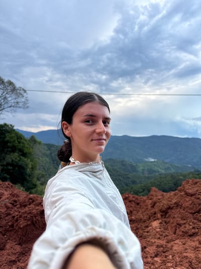 A young woman with dark hair pulled back is taking a selfie in a mountainous, rural landscape. She is wearing a light-colored jacket and a shell necklace. Behind her, rolling green hills and mountains stretch into the distance under a cloudy sky. In the foreground, mounds of reddish-brown soil are visible. The scene appears to be located in or near the city of Nao Sẻ, Vietnam, indicated by the lush, tropical mountain vegetation and the type of soil.