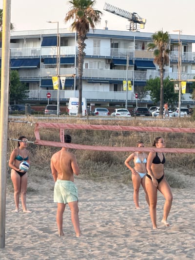 This is a vertical, wide-angle photograph taken at beach level, showing a scene with people playing beach volleyball. The image is oriented such that the subjects and background elements are upside down relative to a standard upright orientation.

In the foreground, the beach volleyball net divides the frame. On one side of the net, a woman in a turquoise bikini is holding a volleyball, appearing to be either preparing to serve or receive. On the other side of the net, a shirtless man is visible, wearing light green shorts. Further in the background, two women in bikinis are playing volleyball. One wears a black bikini and the other a light blue bikini.

Behind the beach volleyball court, a dry, sandy area with sparse vegetation is visible, leading to a tall, multi-story building with balconies. A construction crane is positioned to the left of the building. Palm trees are scattered throughout the scene.

The lighting suggests it is daytime, likely late afternoon or early evening, given the warm tones and soft shadows on the sand. The sky appears bright and clear, indicating fair weather.

Visible text includes "RENT O" on a white van in the background, and smaller yellow signs on the building. There are also markings on the volleyball, including "KIPSTA". The volleyball net itself has a red strap with text that appears to read "AMB Platges".

The overall atmosphere of the image is relaxed and active, capturing a moment of recreational sports on a beach in a coastal urban environment, possibly Castelldefels, Spain, as indicated by the location context.